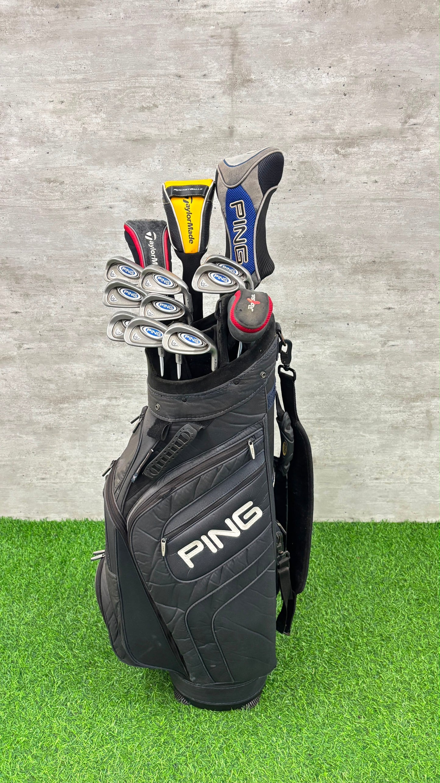 A black PING golf bag filled with golf clubs, standing on artificial green turf against a gray concrete wall.