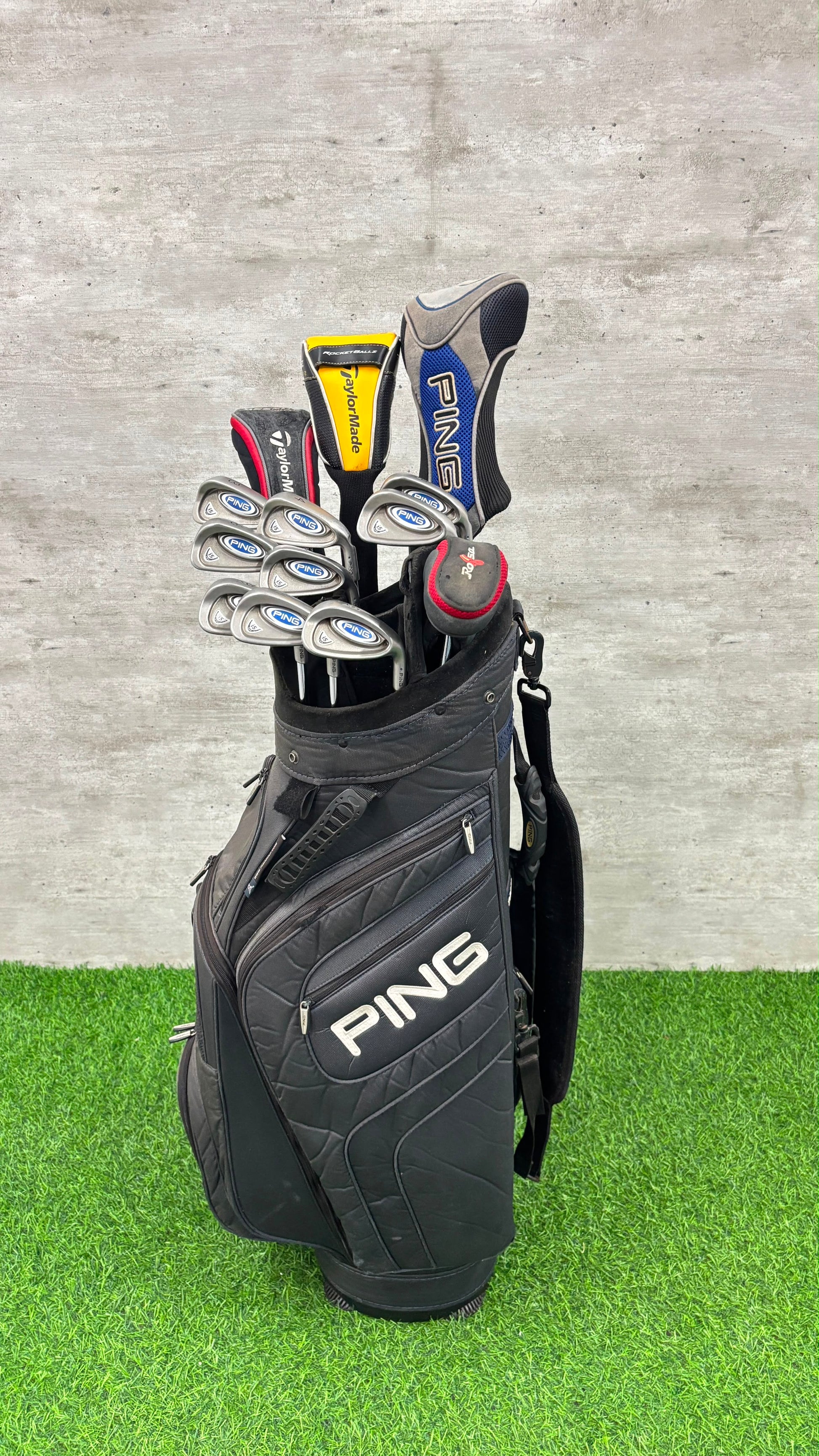 A black PING golf bag filled with golf clubs, standing on artificial green turf against a gray concrete wall.