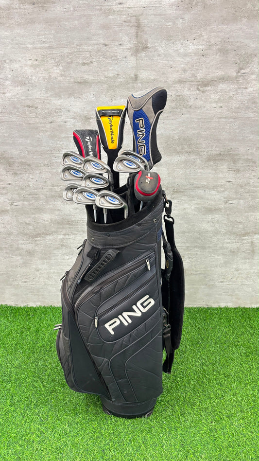 A black PING golf bag filled with golf clubs, standing on artificial green turf against a gray concrete wall.