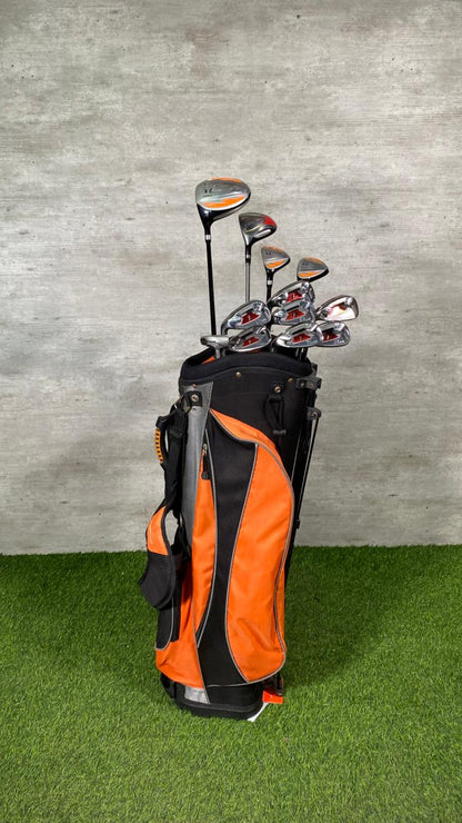 Golf clubs in an orange and black bag on a grassy surface with a gray wall background