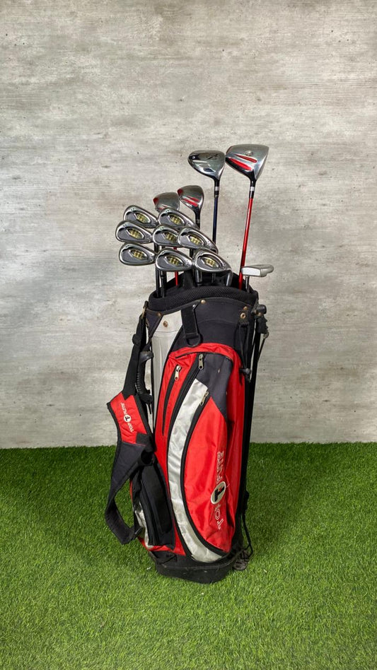 Red and black golf bag with golf clubs on a grassy surface against a gray wall.