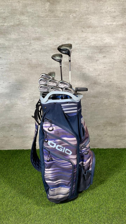 Golf bag with golf clubs on a grassy surface against a concrete wall.