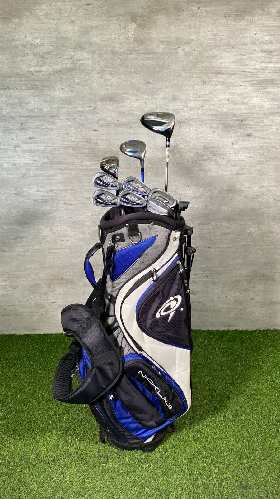 Golf clubs in a bag on a grassy surface with a concrete wall background