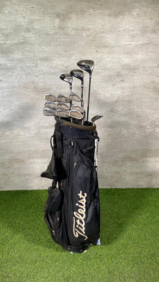 Titleist golf bag with golf clubs on a grassy surface against a concrete wall.