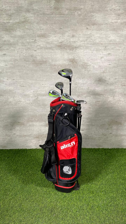 Golf clubs in a red and black bag on a grassy surface with a concrete wall background