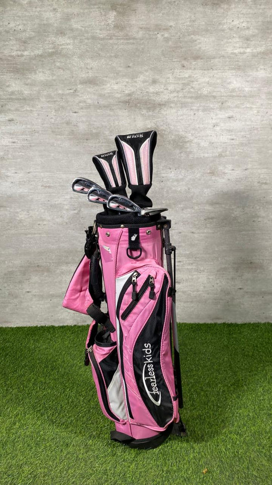 Pink and black golf bag with clubs on a grassy surface against a concrete wall.