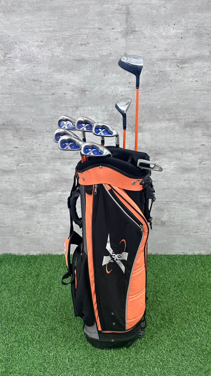 An orange and black golf bag filled with golf clubs, standing on artificial green turf against a gray concrete wall.