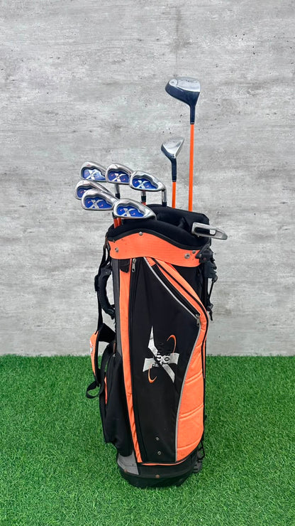 An orange and black golf bag filled with golf clubs, standing on artificial green turf against a gray concrete wall.