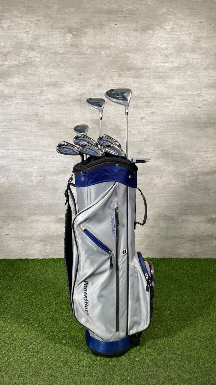 A silver and blue Ping Golfer golf bag filled with golf clubs, standing on artificial green turf against a textured gray wall.