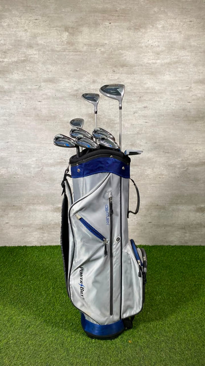 A silver and blue Ping Golfer golf bag filled with golf clubs, standing on artificial green turf against a textured gray wall.