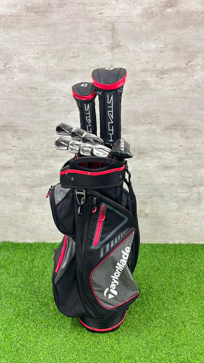 A black TaylorMade golf bag with red and gray accents, containing several TaylorMade golf clubs, set against a gray concrete wall and green artificial turf.