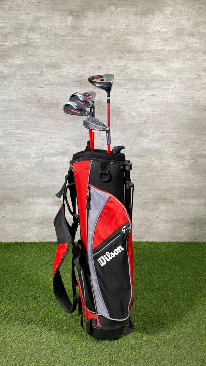 A Wilson golf bag set with several golf clubs, standing on artificial green turf against a textured gray wall.