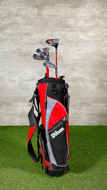 A Wilson golf bag set with several golf clubs, standing on artificial green turf against a textured gray wall.