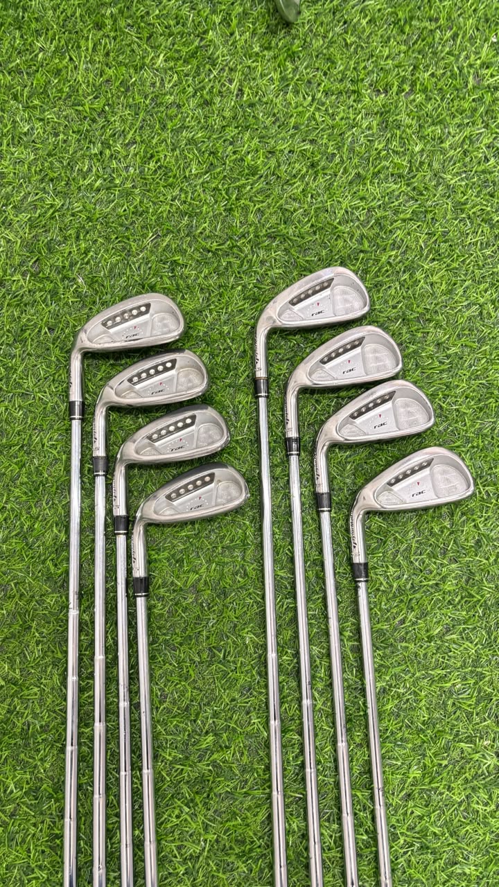 A set of eight golf iron clubs arranged on bright green artificial turf. The clubs have silver metal heads with black and white branding, and long silver shafts with black grip sections.