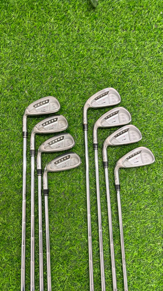 A set of eight golf iron clubs arranged on bright green artificial turf. The clubs have silver metal heads with black and white branding, and long silver shafts with black grip sections.