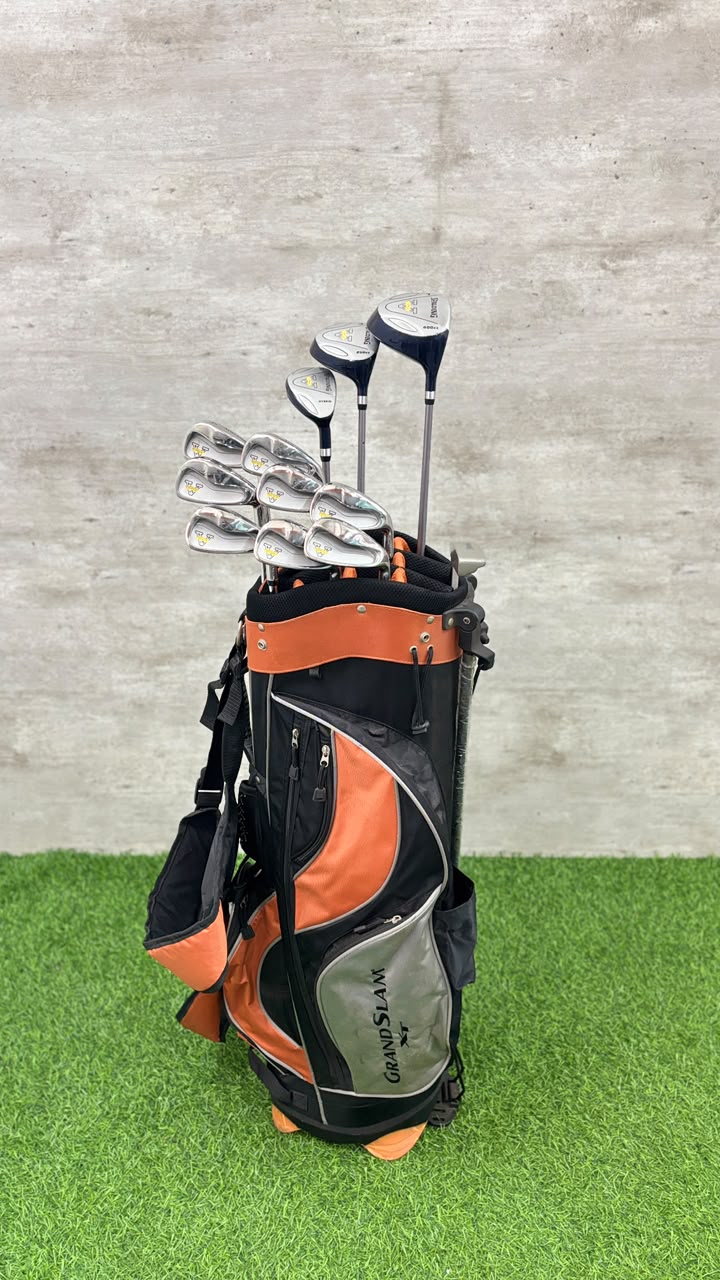 A black and orange Callaway golf bag filled with golf clubs, standing on artificial green turf against a gray textured wall.