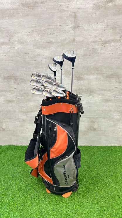 A black and orange Callaway golf bag filled with golf clubs, standing on artificial green turf against a gray textured wall.