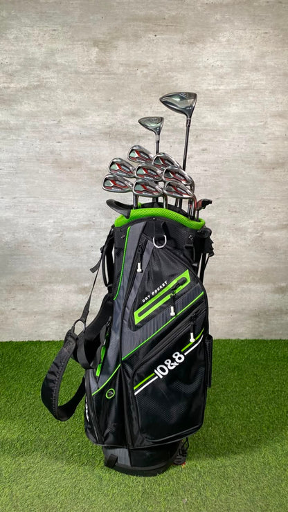 A black and green B&G golf bag filled with golf clubs, standing on artificial grass against a textured gray wall.
