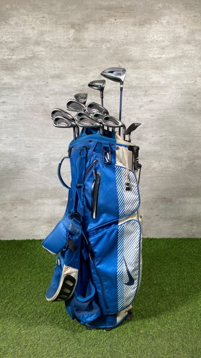 A blue and white golf bag filled with golf clubs, standing on artificial green turf against a textured gray wall.