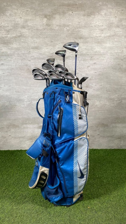 A blue and white golf bag filled with golf clubs, standing on artificial green turf against a textured gray wall.