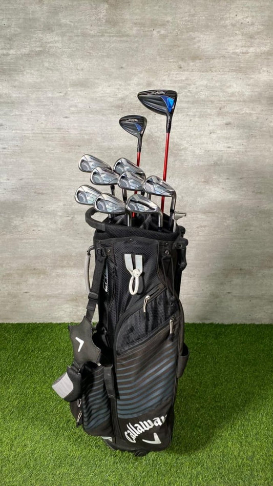 Golf bag with golf clubs on a grassy surface against a concrete wall.
