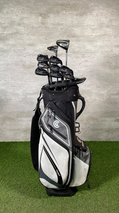 A black and white golf bag filled with golf clubs, standing on artificial green turf against a gray concrete wall.