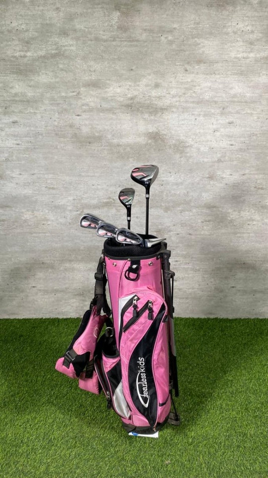 Pink golf bag with clubs on a grassy surface against a concrete wall.