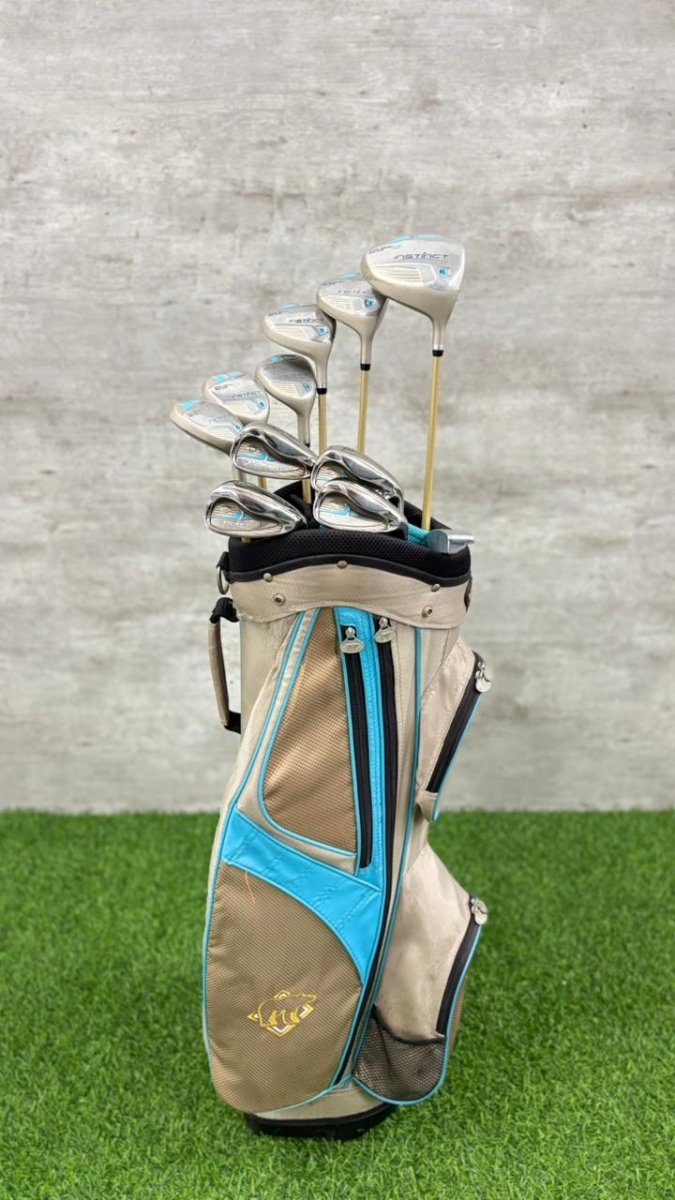 Golf club set in a beige and blue bag on grass with a gray concrete wall background