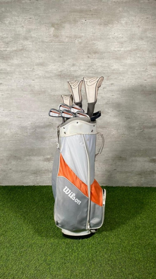 Golf clubs in a gray and orange Wilson bag on grass with a gray wall background