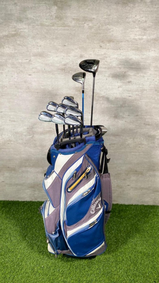 Blue golf bag with golf clubs on a grassy surface against a gray wall.