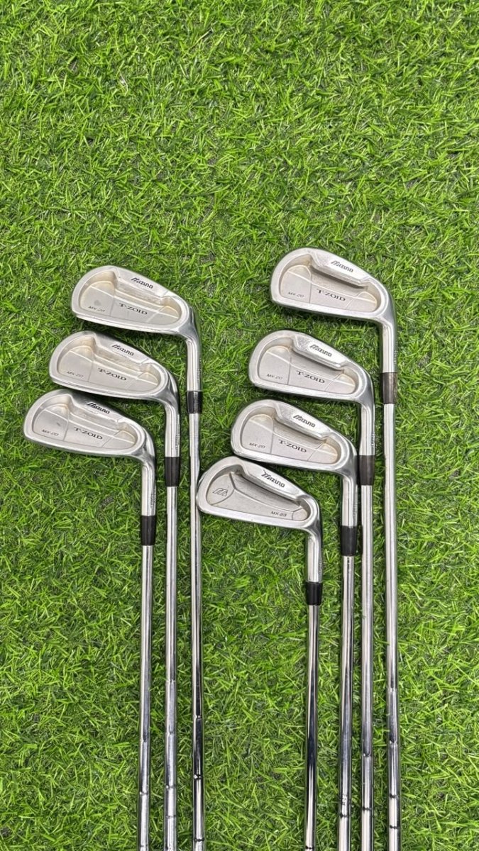 A set of eight golf iron clubs arranged in two groups of four on a green grass surface. The clubs have silver metal heads with black grip sections and silver shafts.
