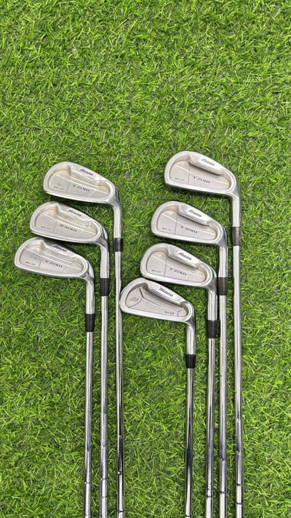 A set of eight golf iron clubs arranged in two groups of four on a green grass surface. The clubs have silver metal heads with black grip sections and silver shafts.