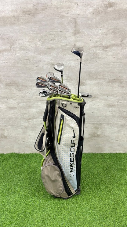 A Nike golf bag filled with golf clubs, standing on artificial green turf against a textured gray wall. The bag is white and gray with black and neon yellow-green accents, and the Nike swoosh and 'NIKE GOLF' branding are clearly visible.