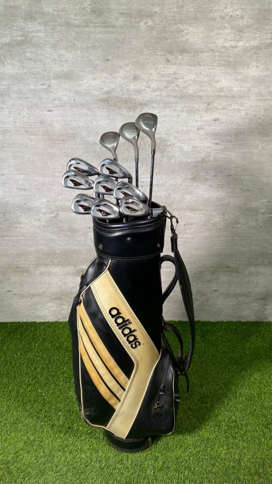 Golf clubs in a black and gold Adidas bag on a grassy surface with a concrete wall background