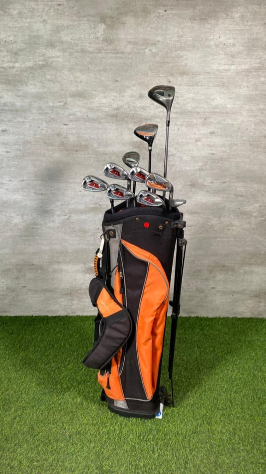 Golf clubs in an orange and black bag on a grassy surface with a gray concrete wall background