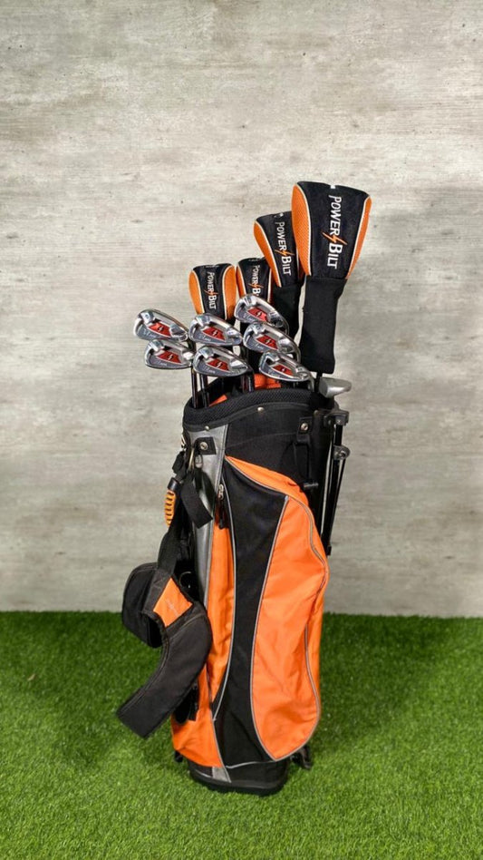 Golf clubs in an orange and black bag on a grassy surface with a concrete wall background