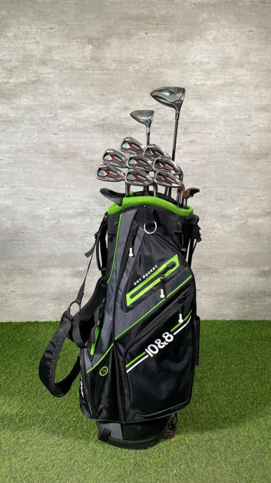 A black and green B&G golf bag filled with golf clubs, standing on artificial grass against a textured gray wall.