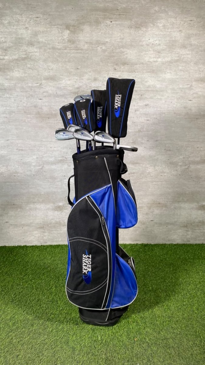 A black and blue golf bag filled with golf clubs, standing on artificial green turf against a gray textured wall. The bag and headcovers feature the brand name 'STOCK' in white.